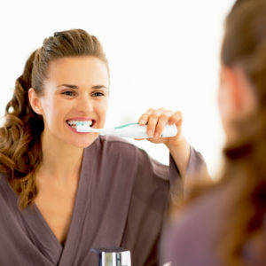 Our Grosse Pointe dentist provides insight on the pros and cons of electric toothbrushes. In this image, a woman smiles as she brushes her teeth in front of a mirror.